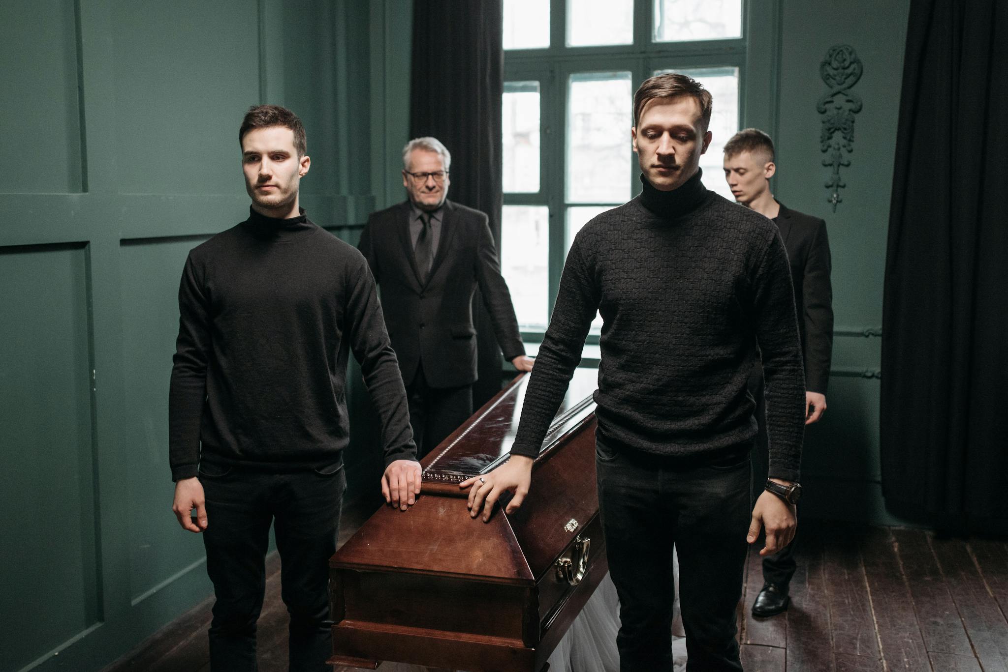A group of men stands solemnly around a coffin in a dimly lit room, reflecting sorrow and reverence.